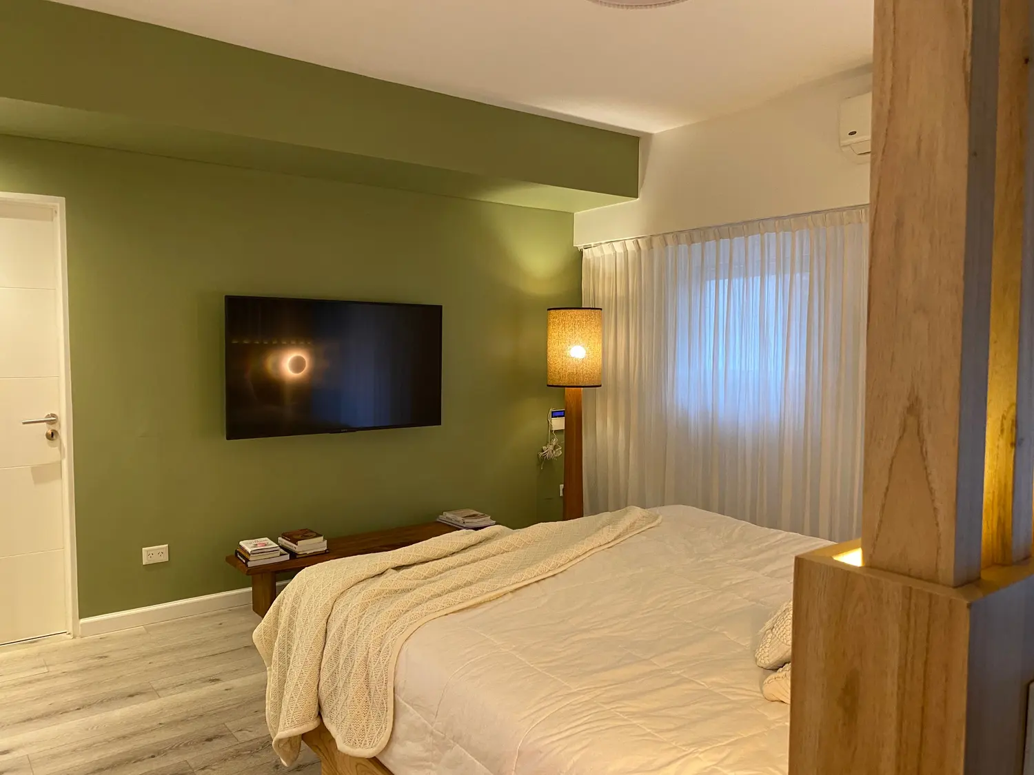 Dormitorio con pared verde, televisor y lámpara de pie; adelante, la cama y una columna de madera