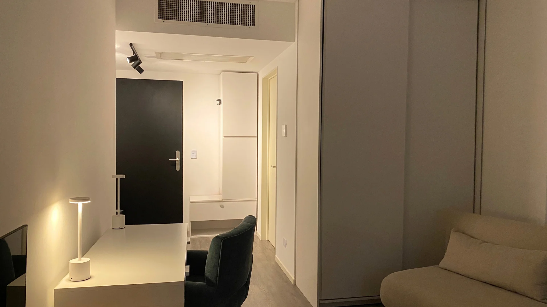 Escritorio con lámpara, la puerta de entrada negra y el placard blanco con un sillón