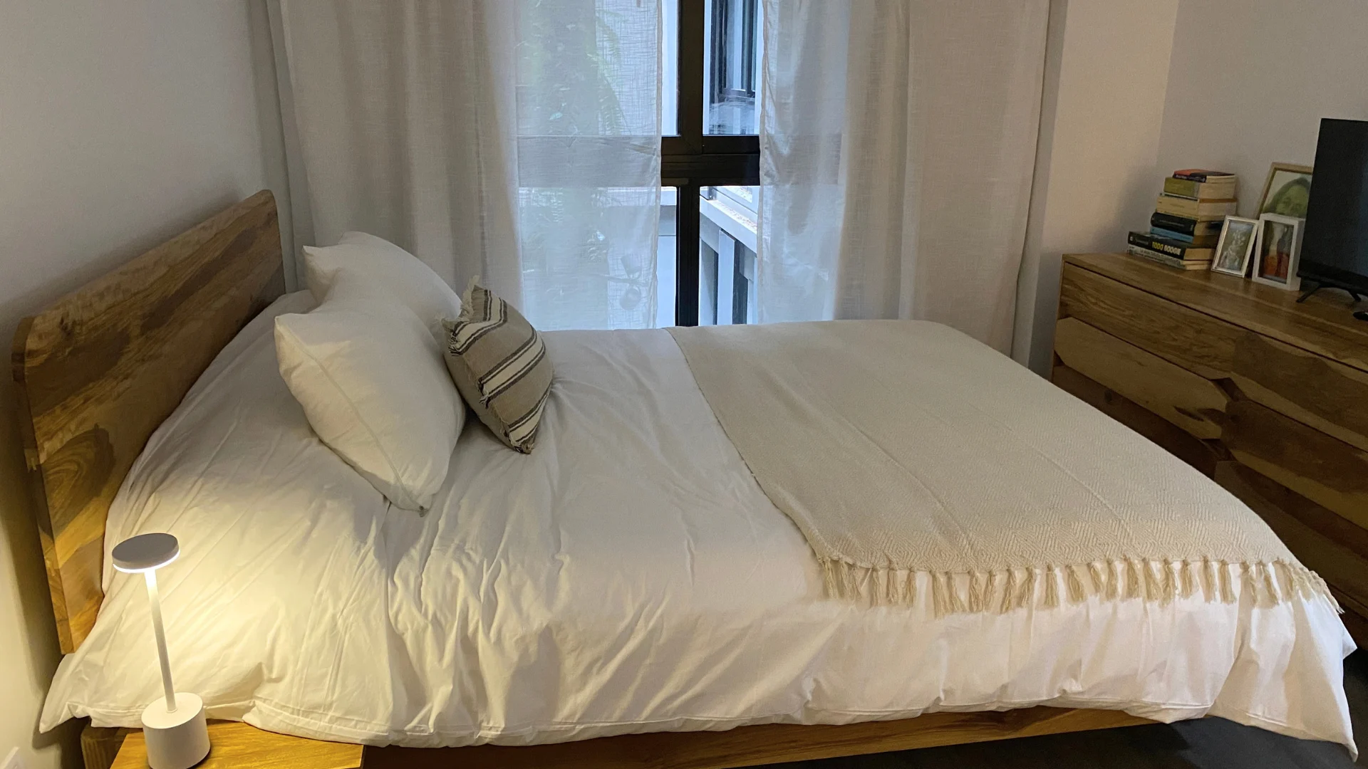 Cama de madera con ropa de cama blanca y almohadones a rayas, junto a la ventana